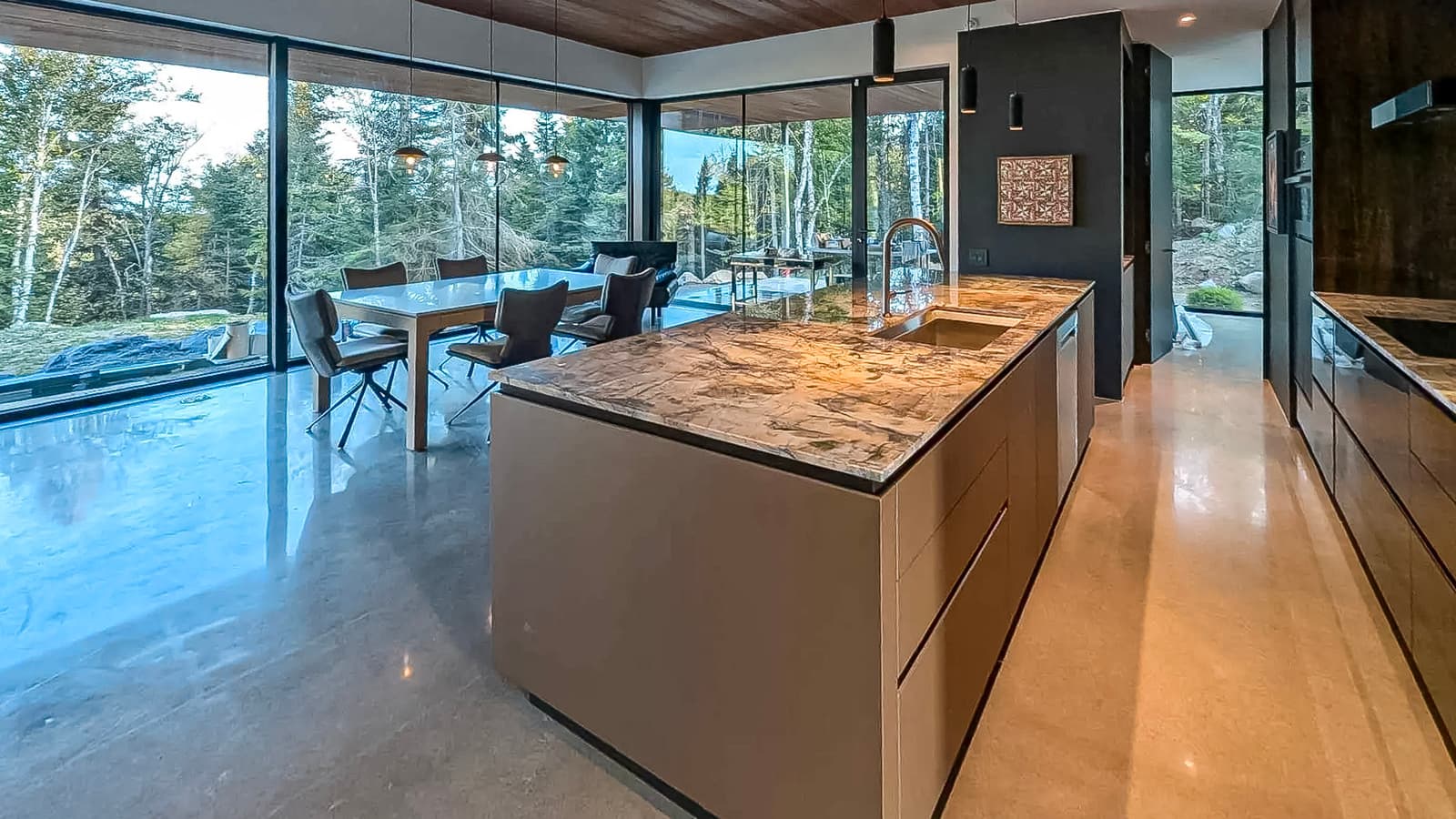 Modern kitchen with a marble island, large glass windows overlooking the forest, wooden ceiling, and open dining area.