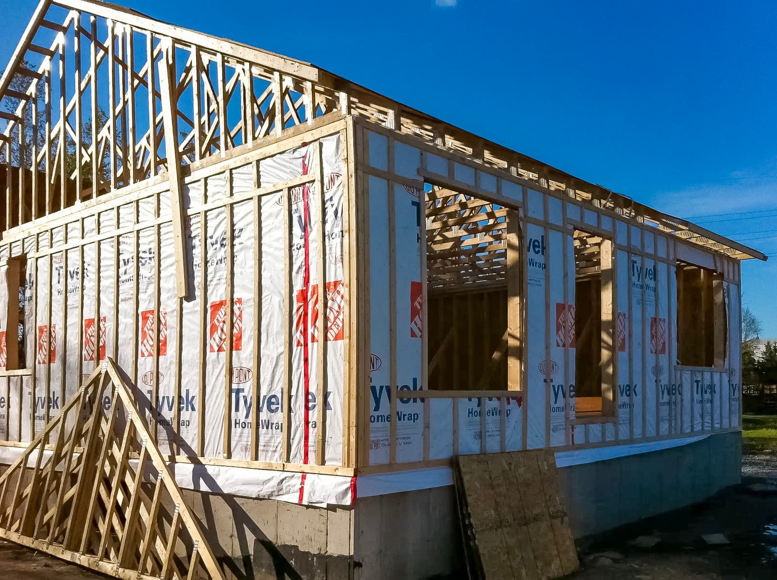 Wooden house structure under construction with Tyvek weather wrap, visible framing, and concrete foundation under a blue sky.