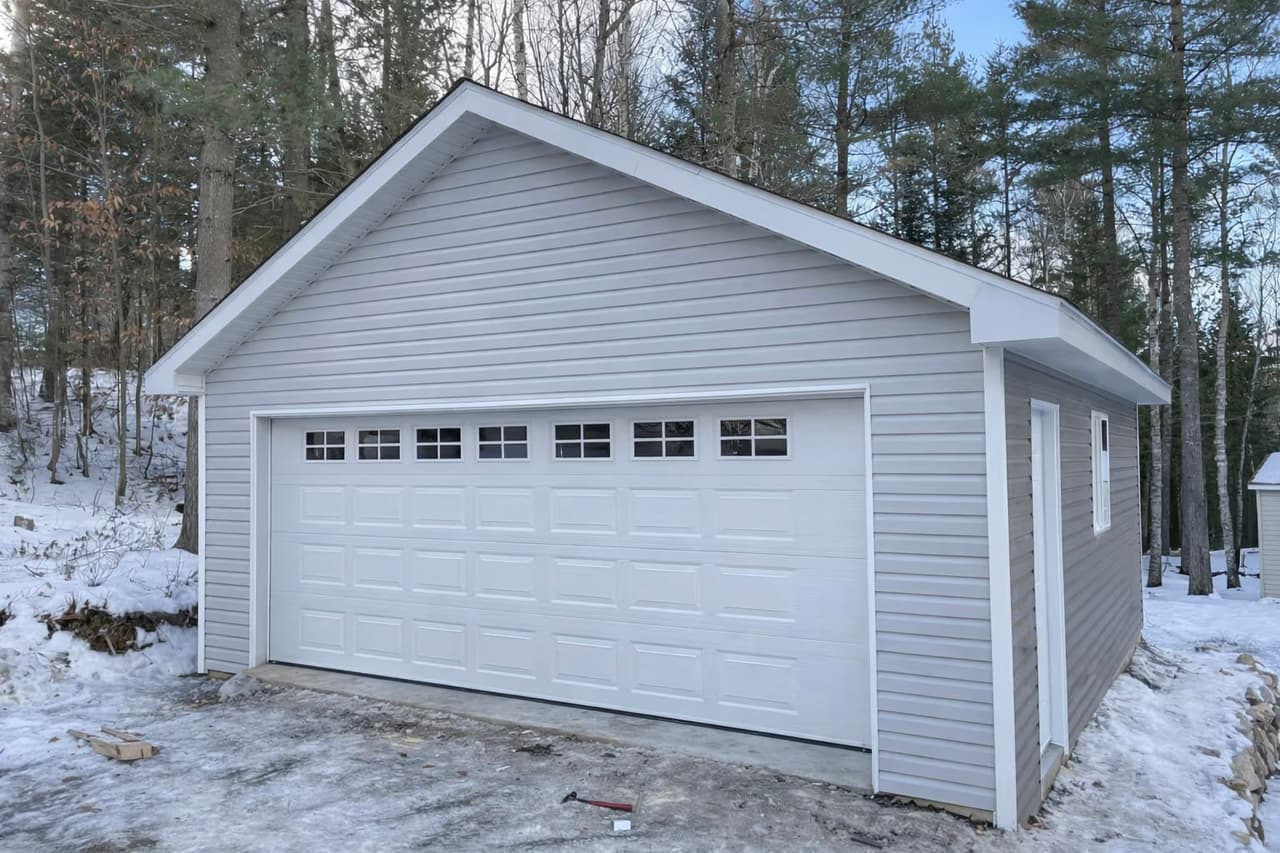 Modern detached garage with gray vinyl siding, large white garage door, white soffits and fascia, gable roof in a snowy wooded setting.