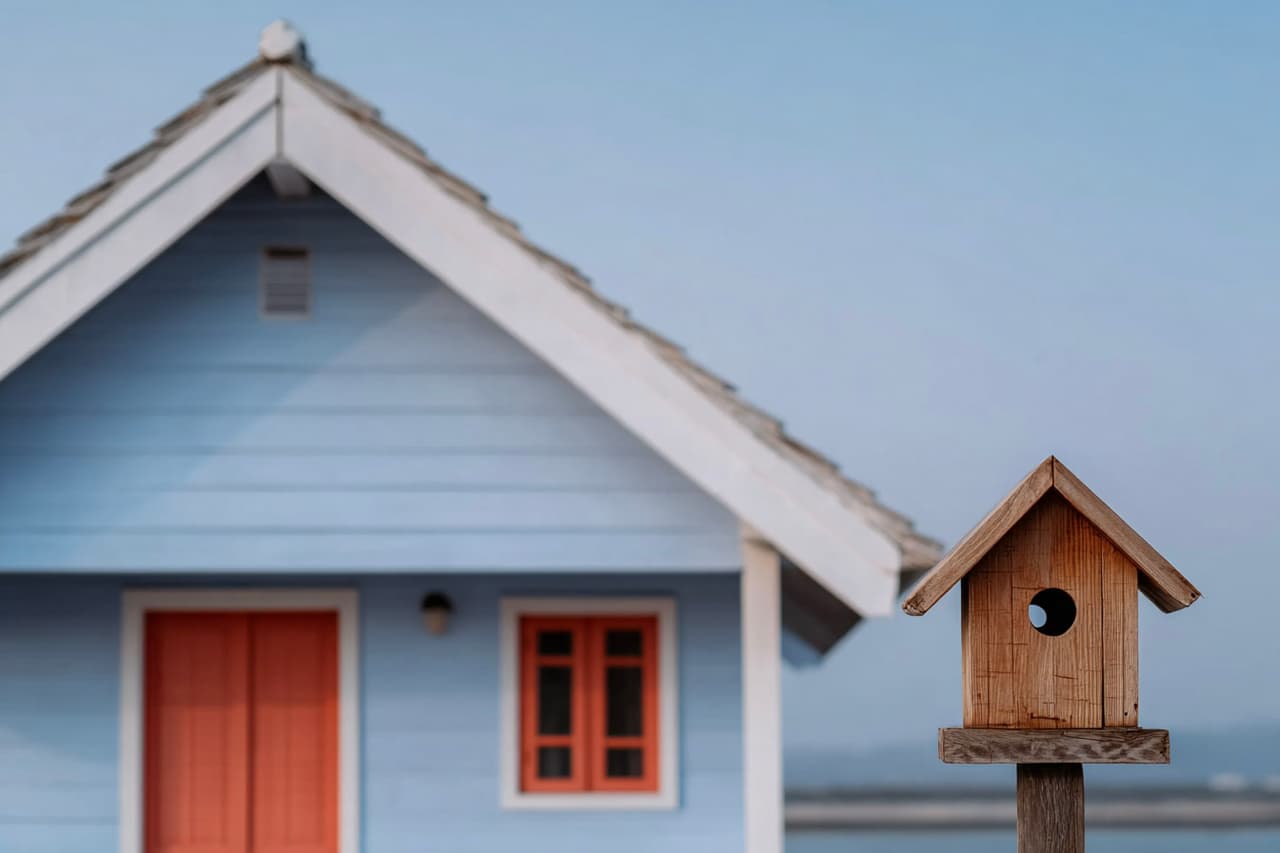 Wooden birdhouse mounted on a post in front of a blue house with red door and window, pitched roof and softly blurred outdoor background.