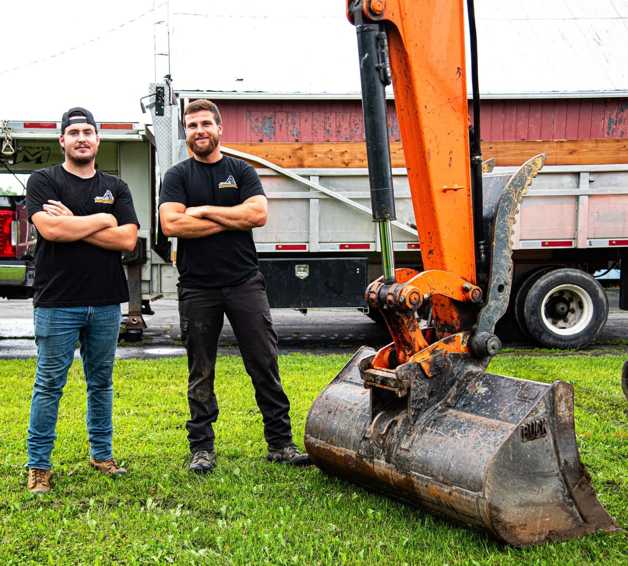 Two construction workers proudly posing in front of an orange excavator and a work truck on a grassy field