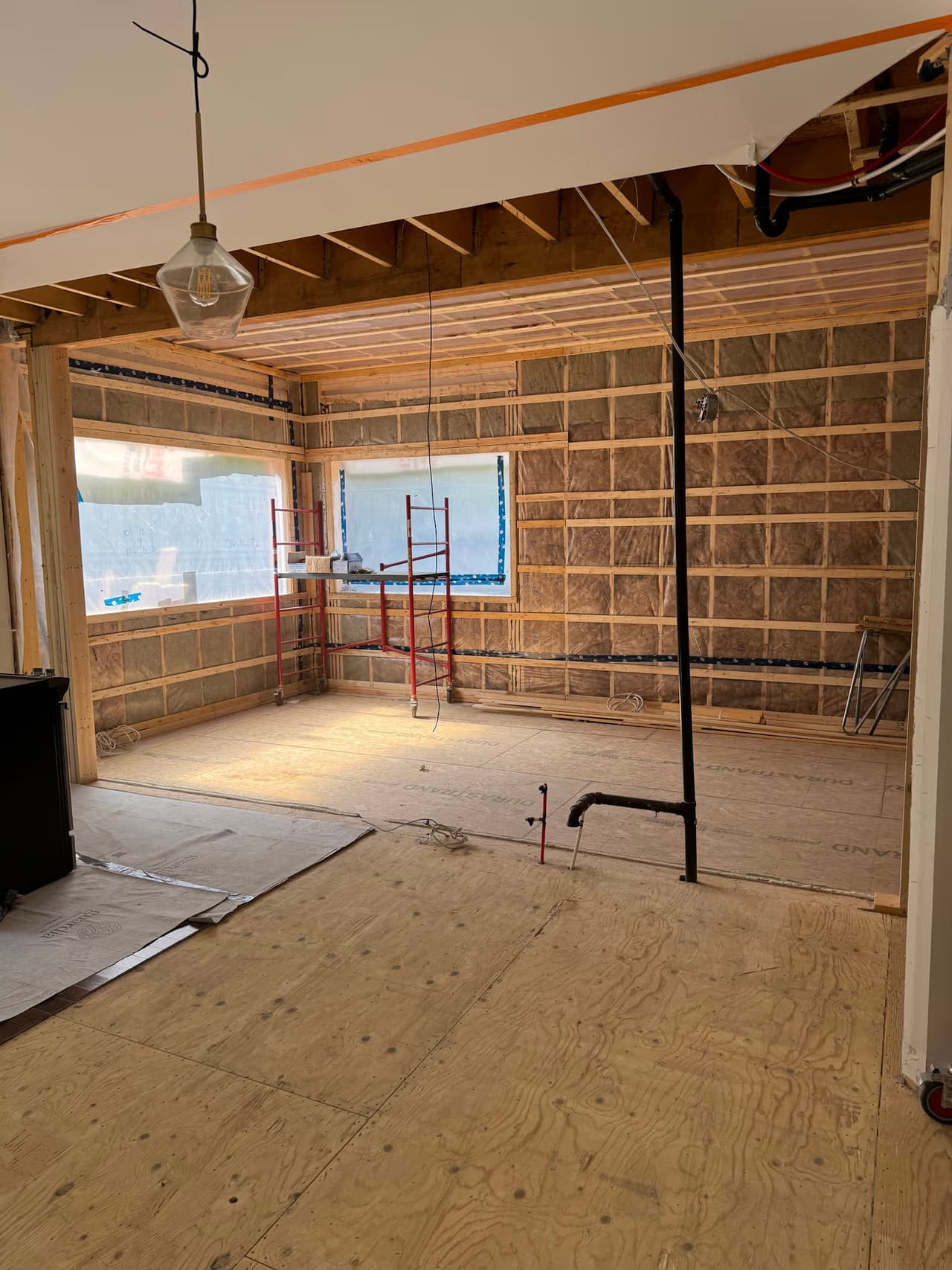 Room under renovation with exposed insulation, wood framing, wiring, plumbing and plywood subfloor.