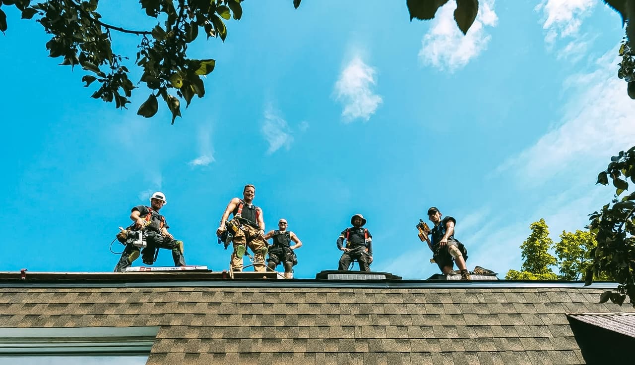 Team of roofers working on a roof under a blue sky, wearing harnesses and construction tools.