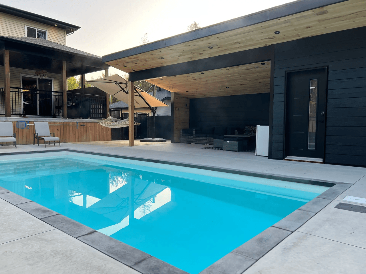 Modern backyard with inground pool, concrete slab terrace, black pavilion with wood ceiling, recessed lighting, exterior door and covered lounge area