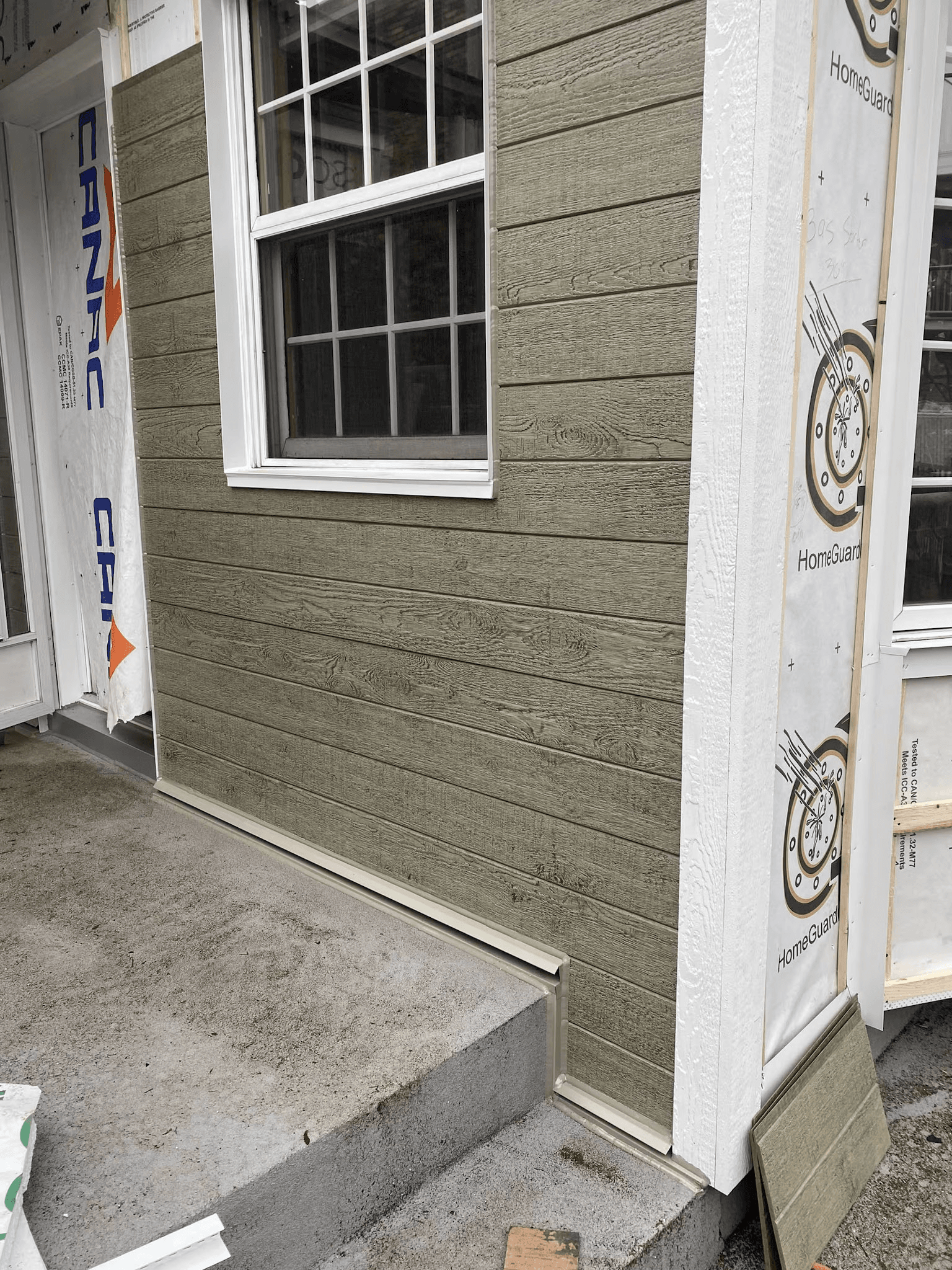 Installation of horizontal wood-textured exterior siding around a window, with weather barrier, white trim and concrete steps on a house under renovation.