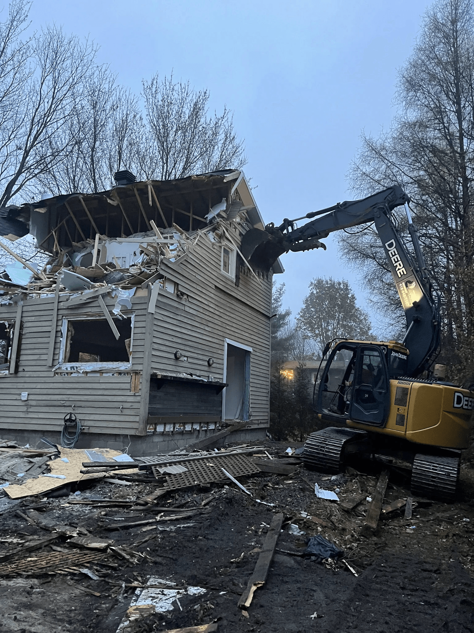 Mechanical demolition of a residential house using a tracked excavator, with partially demolished roof and walls and construction debris on the ground.