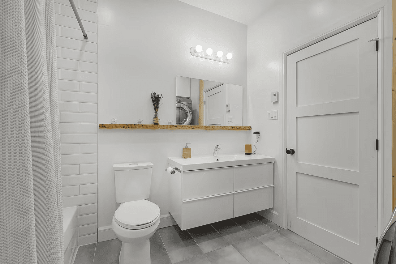 Bright modern bathroom with white floating vanity, wall mirror, toilet, shower with white tile and gray flooring.
