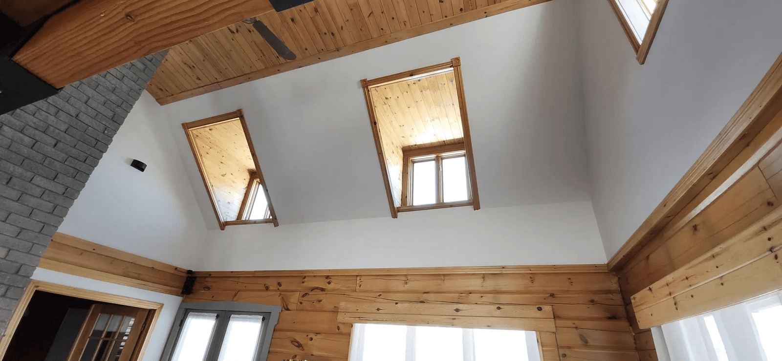 Residential interior featuring a natural wood cathedral ceiling, exposed timber beams, high windows, white walls and a gray brick fireplace in a contemporary rustic style.