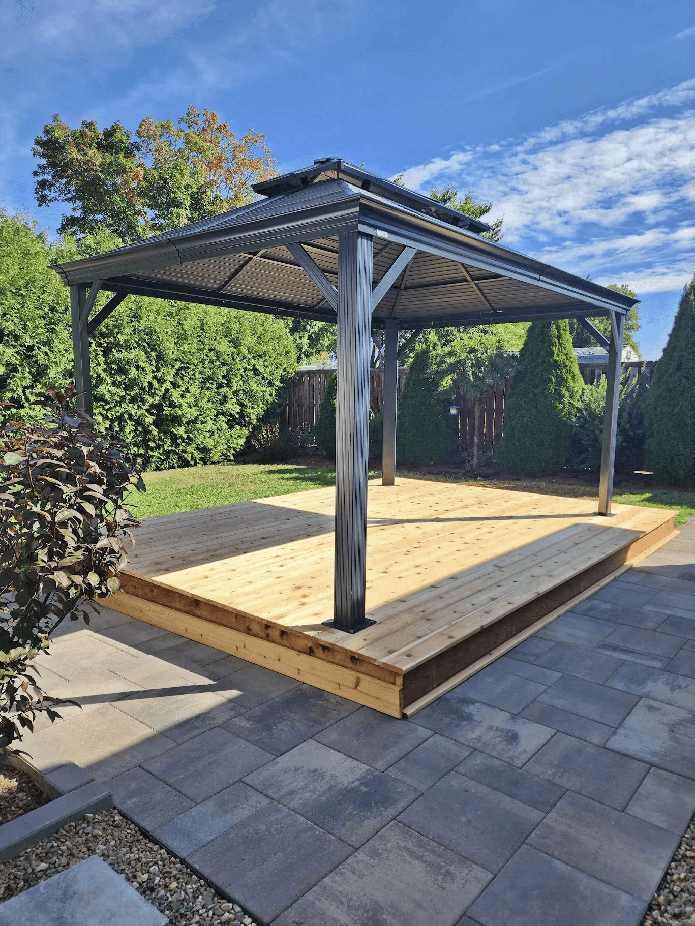 Modern dark gray metal gazebo installed on a raised natural wood deck, surrounded by interlocking pavers and hedges in a landscaped residential backyard.