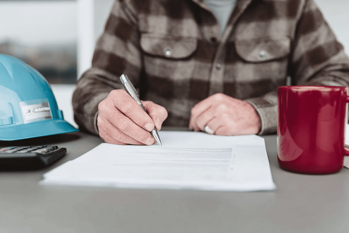 Construction contractor completing and signing administrative documents at a table with a hard hat, calculator and coffee mug