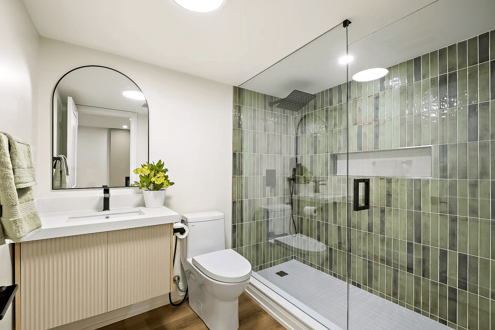 Modern bathroom with glass shower, green tiles, light wood vanity, arched mirror, and stylish accessories.