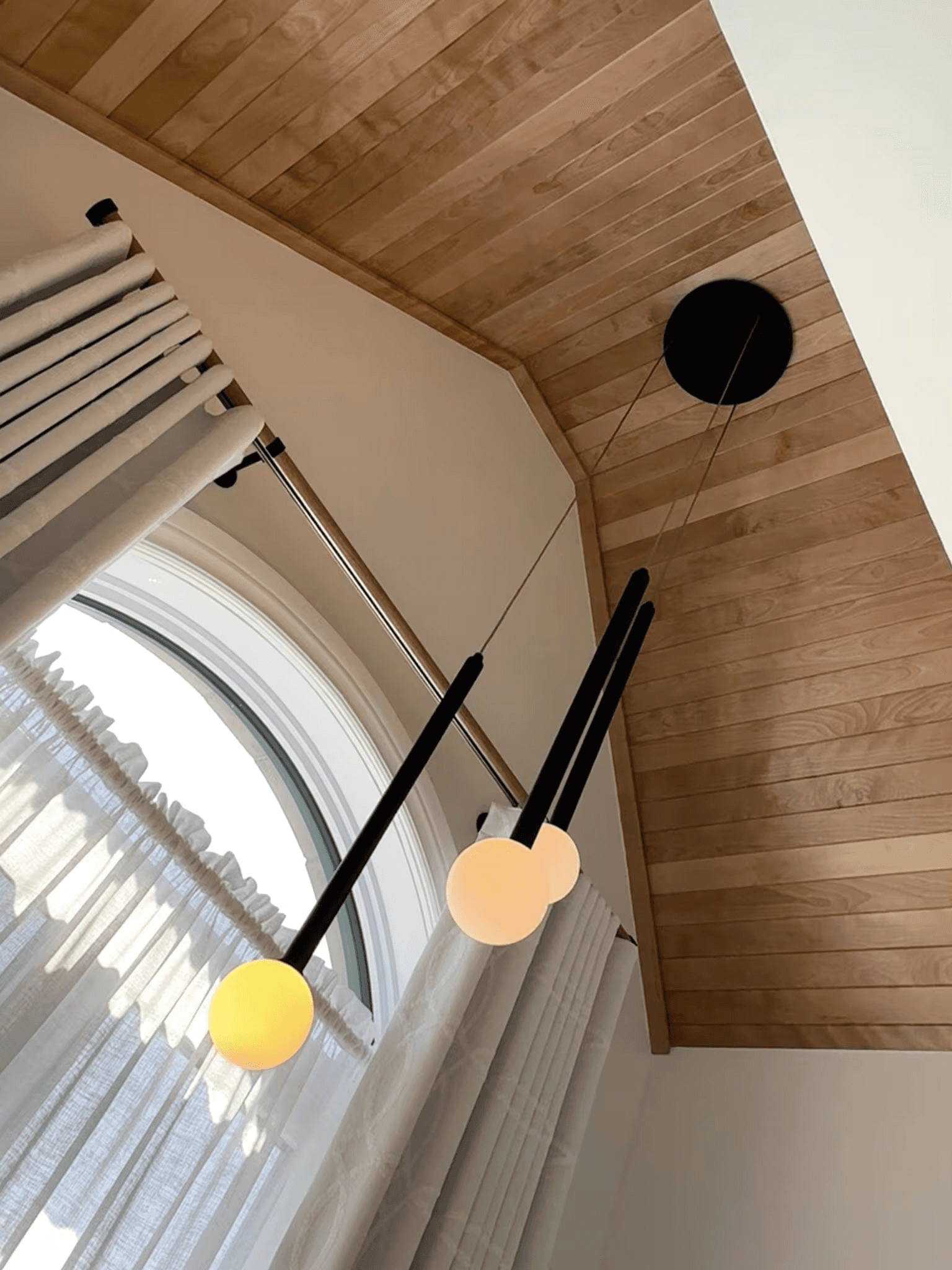 Natural wood-paneled cathedral ceiling with modern black pendant light featuring three illuminated globes, arched window, white curtains and contemporary interior finishes.