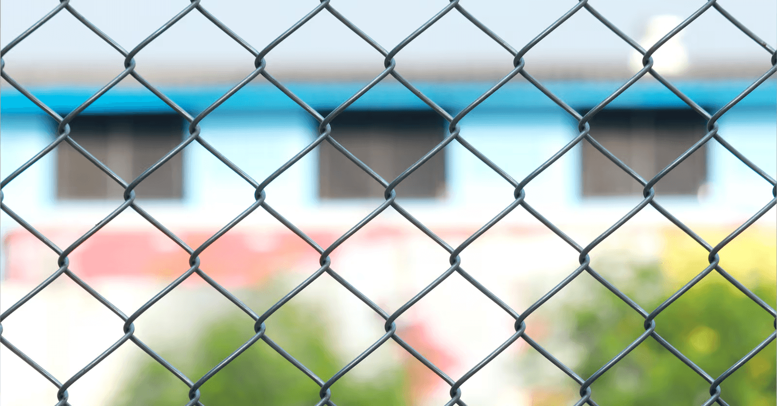 Close-up of a metal chain-link fence with a diamond-shaped wire mesh pattern and a blurred building in the background.