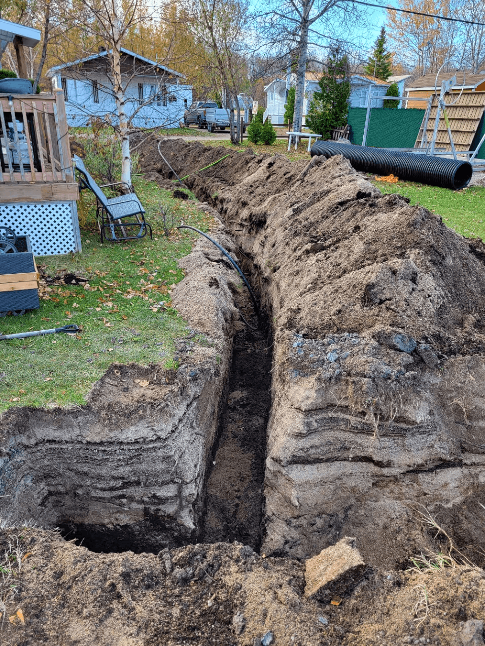 Residential excavation work in a backyard featuring a long trench dug into the ground, black drainage pipes, piles of excavated soil and corrugated drainage tubing, illustrating the preparation of an underground drainage system on a residential property
