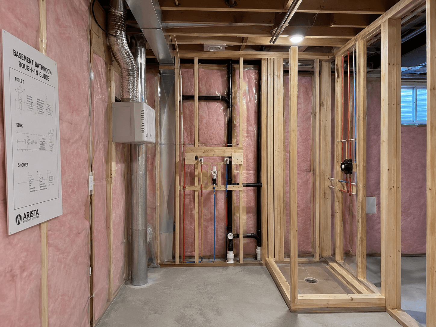 Basement bathroom under construction with wood framing, fiberglass insulation, exposed plumbing, ventilation ducts, and a posted rough-in layout plan