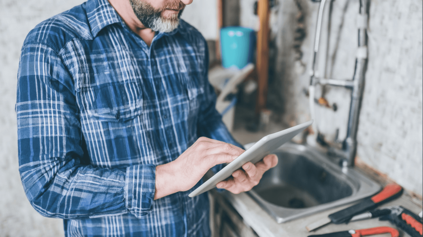 Professional plumber using a digital tablet in front of a sink with exposed piping and plumbing tools.