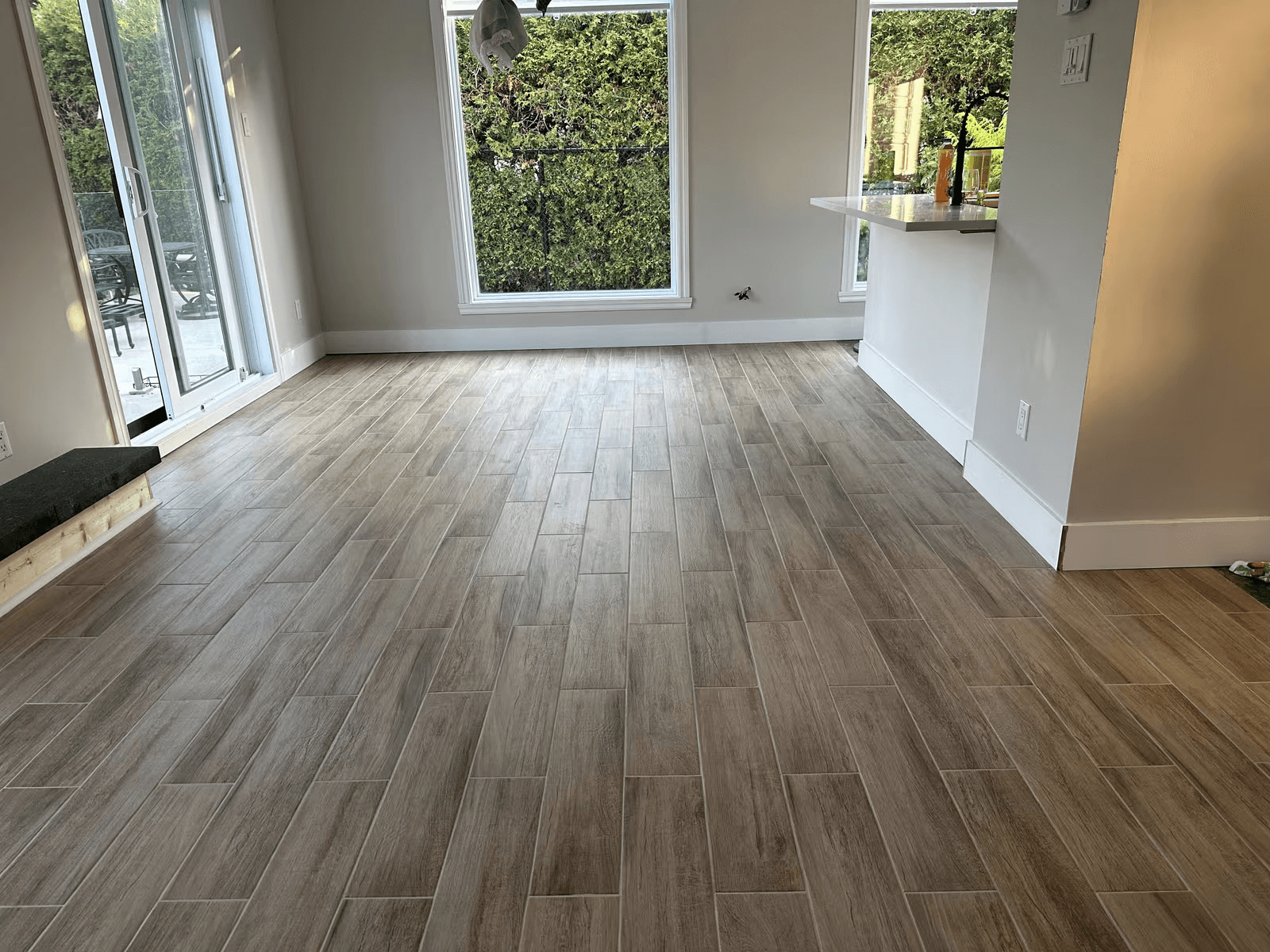 Gray-brown wood-look ceramic tile flooring installed in a bright residential interior featuring large windows, a glass patio door, white walls and a modern kitchen countertop.