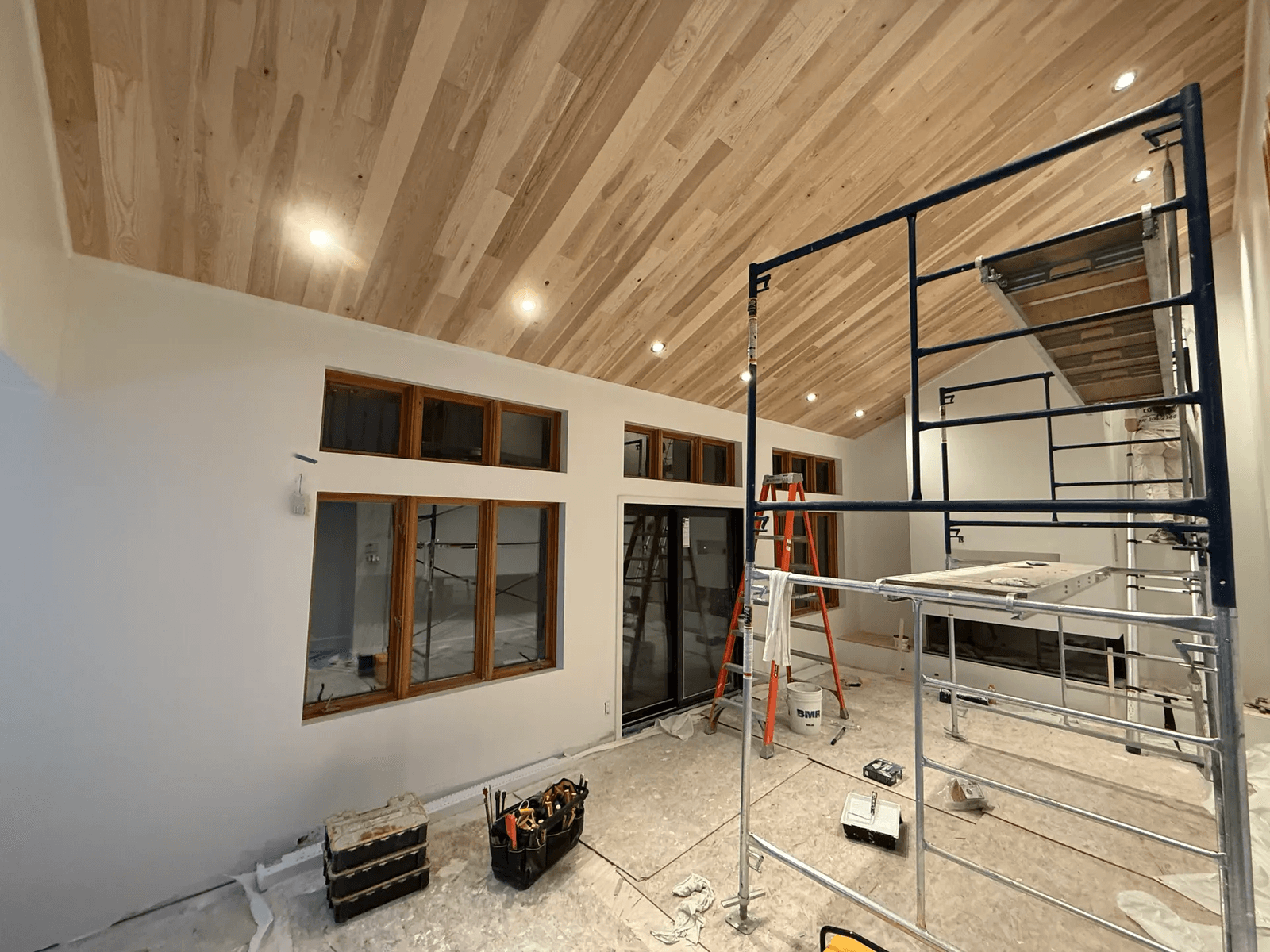  Interior renovation in progress with a cathedral ceiling clad in wood slats, recessed lighting, large windows, glass doors, scaffolding, and construction tools