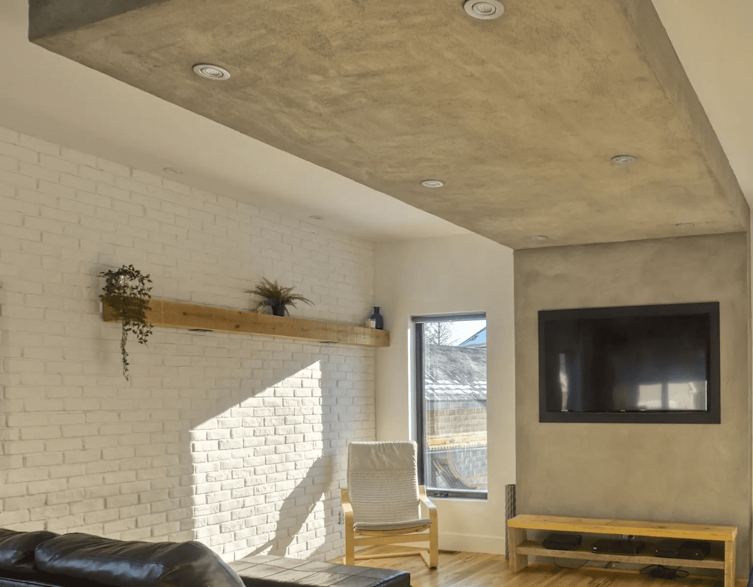 Contemporary living room featuring gray concrete-effect ceiling and wall, recessed lighting, white brick accent wall, natural wood floating shelf, black built-in fireplace and light wood flooring.
