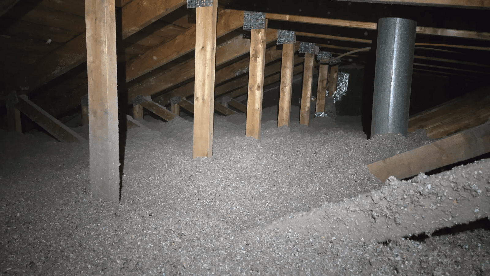 Residential attic with gray blown-in insulation distributed between wooden roof framing, support posts, roof trusses and metal ventilation duct, illustrating attic thermal insulation work to improve home energy efficiency and reduce heat loss