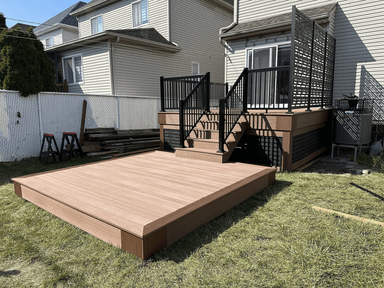 Raised brown composite deck with black aluminum railings, central stairs, privacy screens and matching lower platform in a residential backyard.