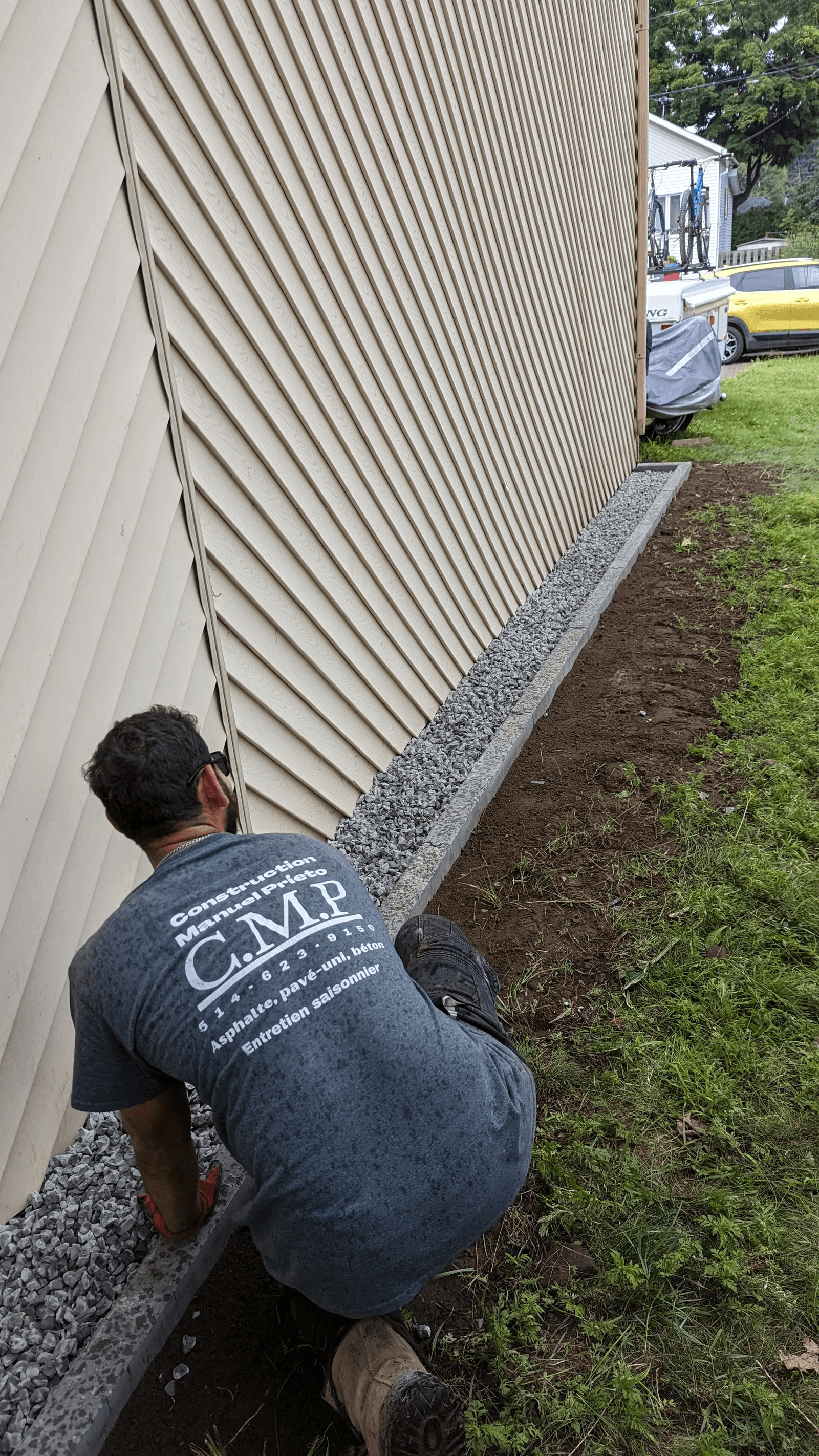 Residential exterior landscaping work featuring a contractor kneeling to install a concrete edging along the beige vinyl siding of a house, outlining a strip of gray crushed stone designed for drainage and landscaping around the foundation