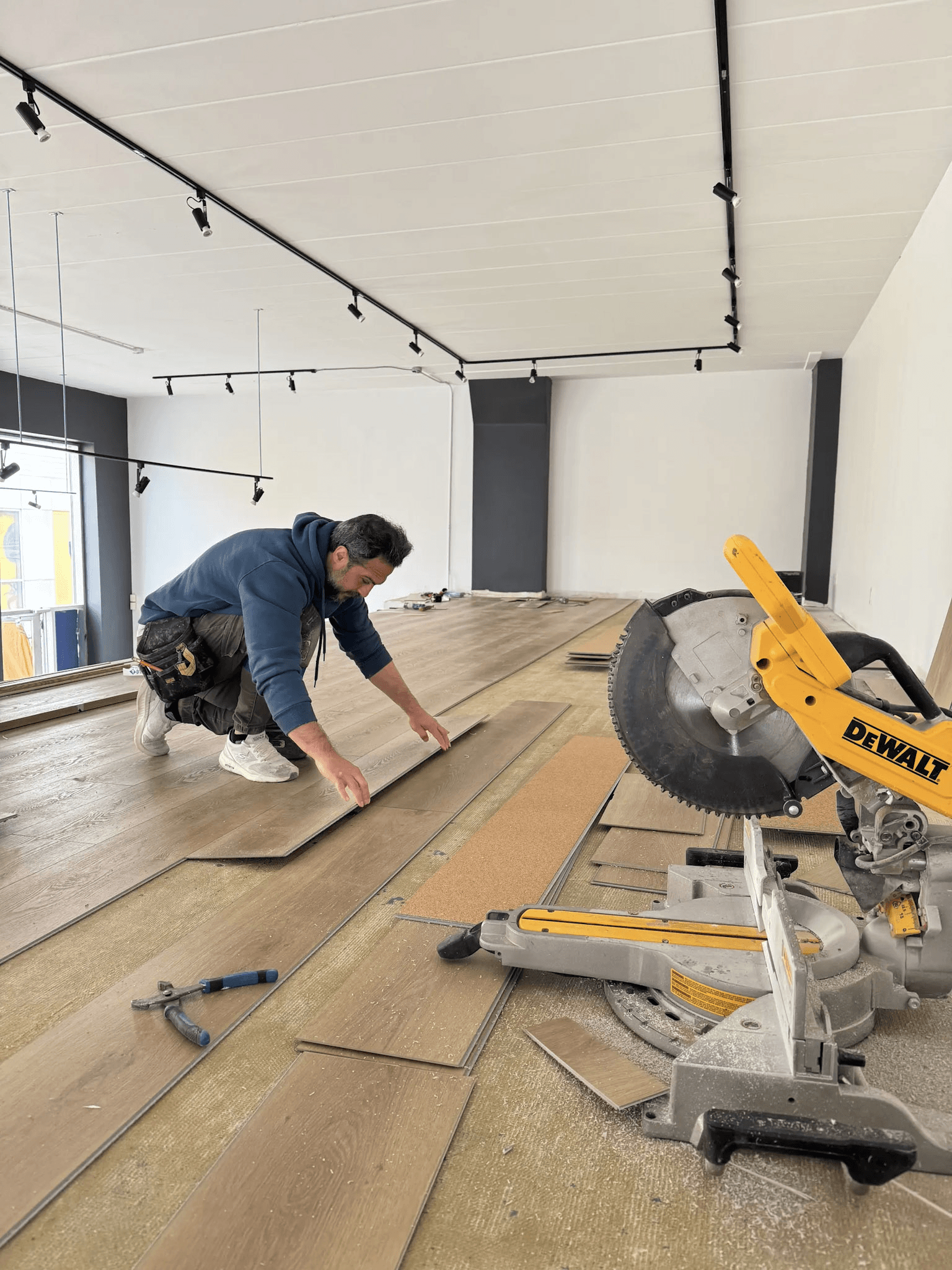 Wood-look flooring installation in a commercial space under renovation, featuring a flooring installer kneeling to position floor planks, a miter saw and ceiling track lighting.
