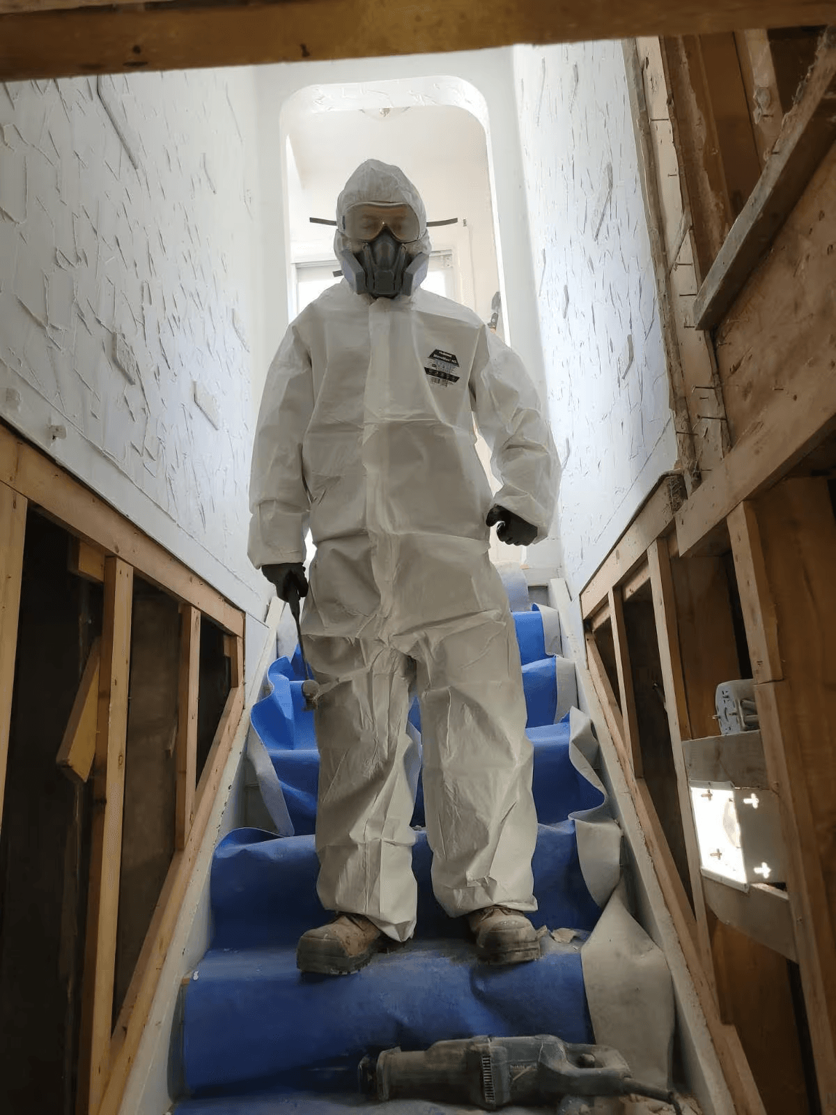 Decontamination technician wearing a full white protective coverall, filtered respirator, safety goggles and gloves, standing on blue protective sheeting covering a staircase at an interior renovation site, illustrating specialized asbestos abatement procedures and hazardous material removal