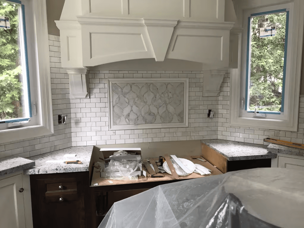 Kitchen under renovation with a custom white decorative range hood, subway tile backsplash, stone countertops, new windows, and cabinetry being installed.