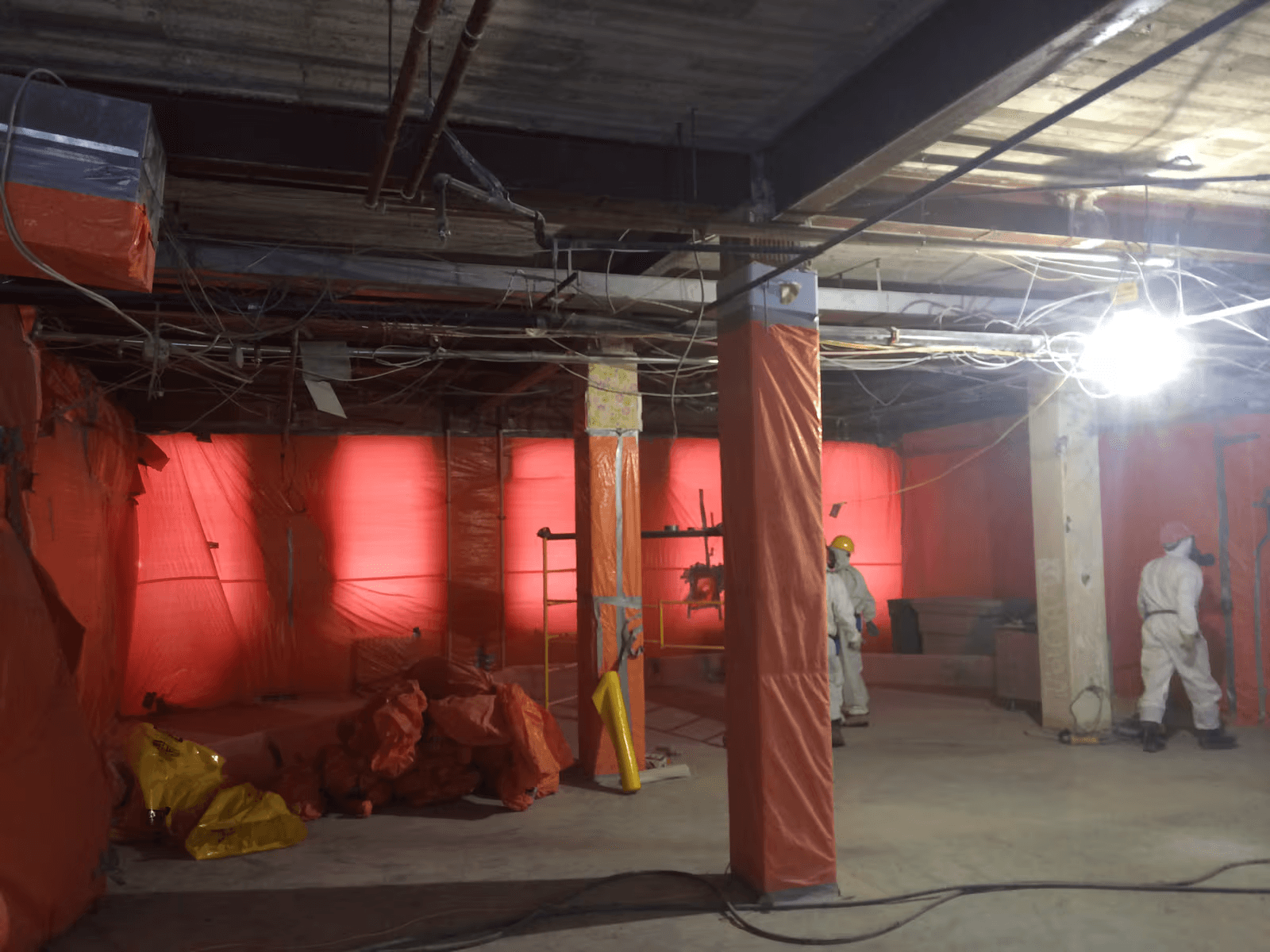 Interior decontamination worksite in a commercial building with two workers wearing white protective coveralls, walls and columns covered with orange plastic containment sheeting, exposed ceiling wiring and waste bags, illustrating specialized containment and hazardous material removal operations