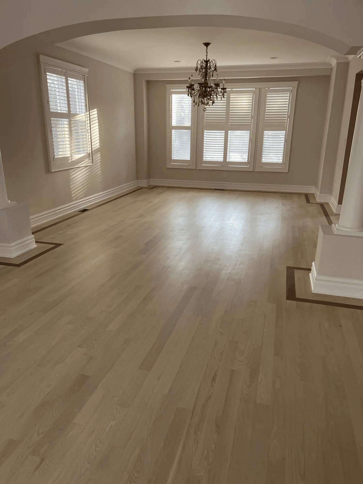 Elegant residential dining room featuring wide-plank light wood flooring, dark wood decorative borders, a classic chandelier, white plantation shutters, decorative moldings and an interior archway.