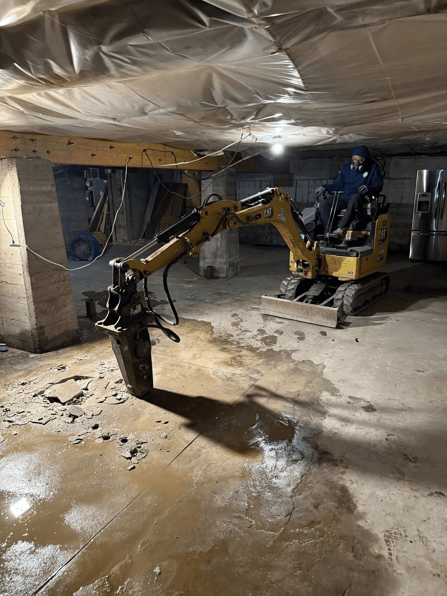 Compact excavator equipped with a hydraulic breaker demolishing a concrete slab inside a residential basement under renovation.