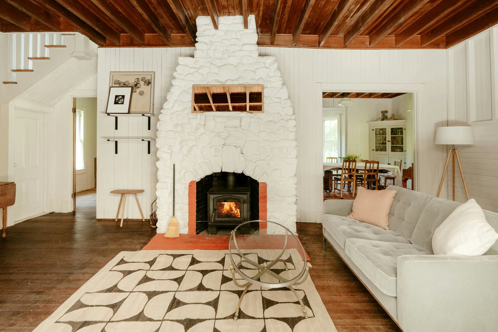 Bright rustic-style living room with a white stone fireplace, lit wood-burning stove, exposed-beam ceiling, hardwood flooring, and contemporary furniture.