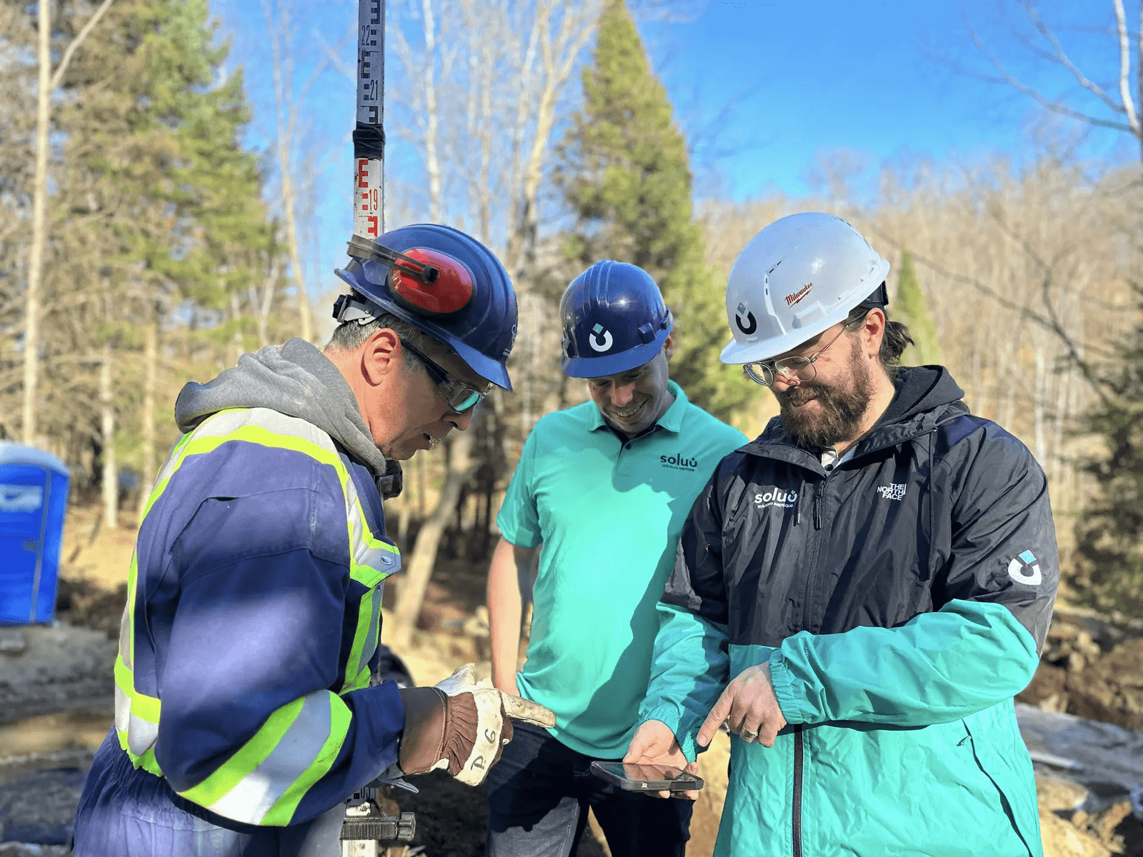 Three construction professionals wearing safety hard hats review information together on a mobile phone at an outdoor wooded job site.