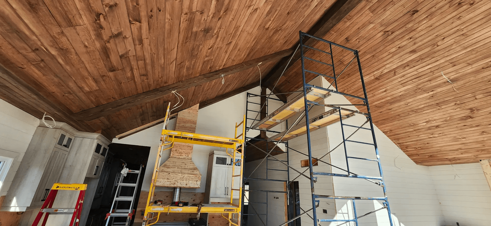 Interior renovation in progress with a large wood cathedral ceiling, exposed beams, white walls, scaffolding, and visible electrical wiring