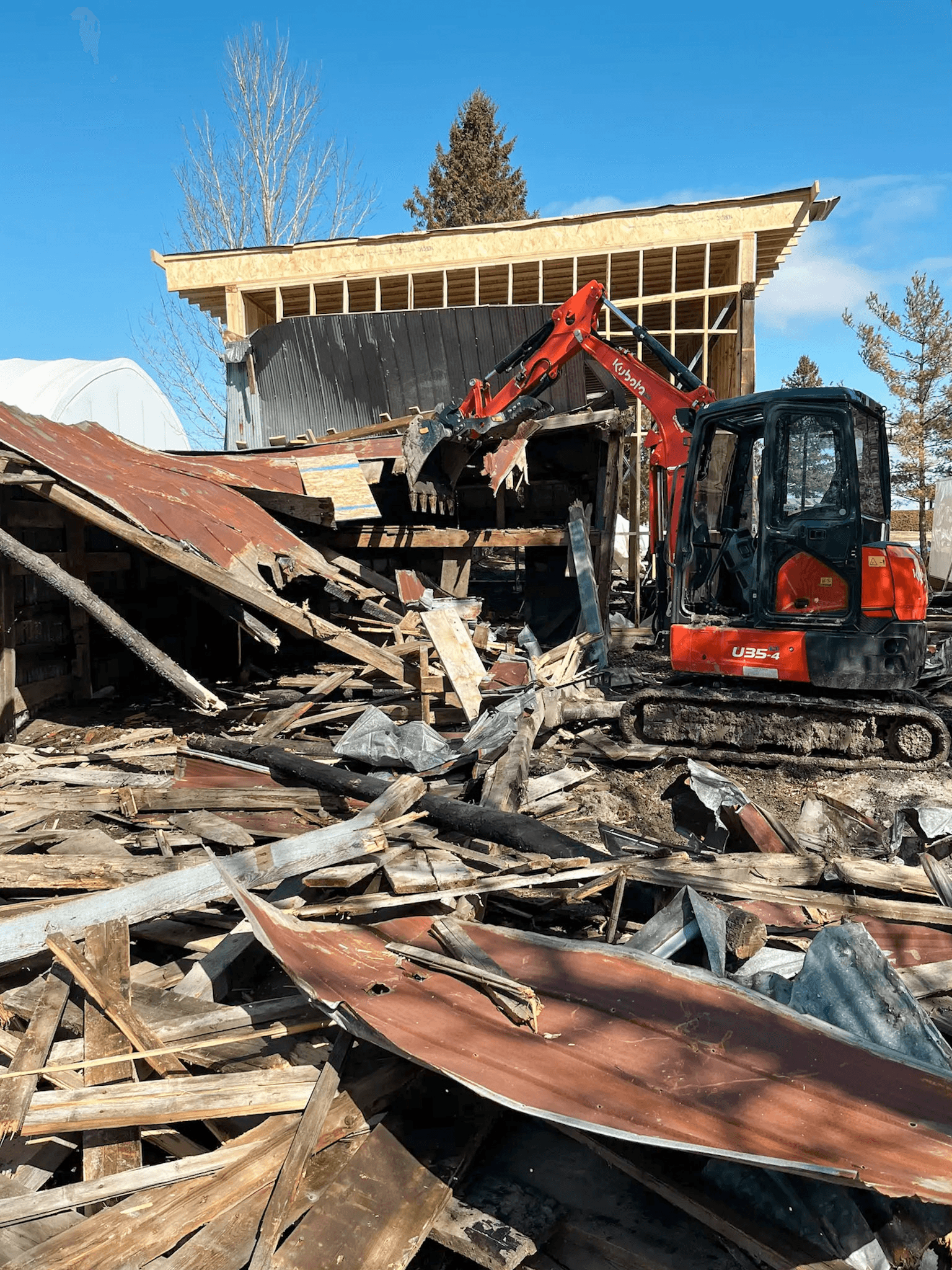 Demolition site with excavator tearing down old wooden structure in front of a new building under construction.