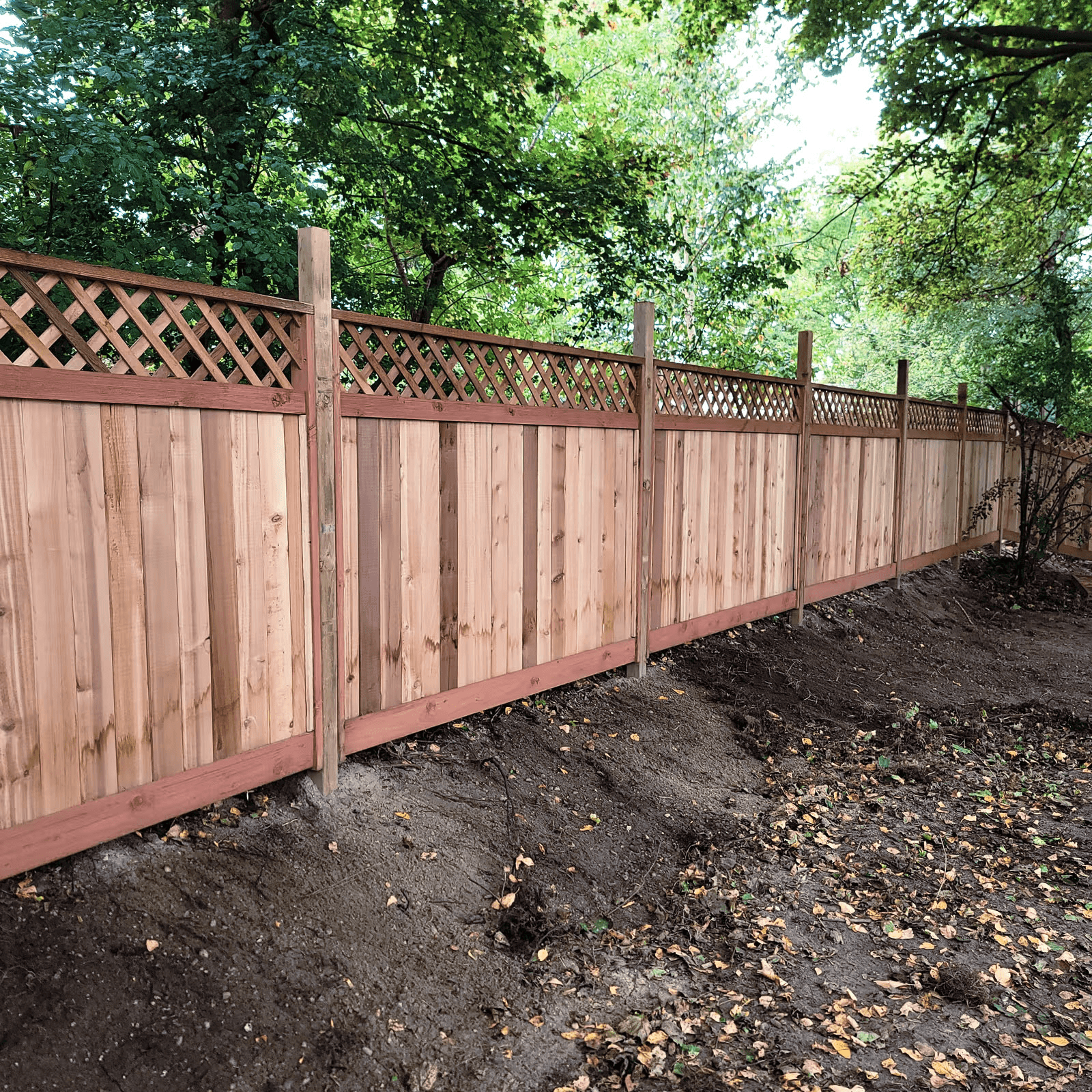Installation of a natural wood privacy fence with vertical panels, decorative diamond lattice and support posts, enclosing a wooded residential property while providing privacy, security and harmonious landscaping.