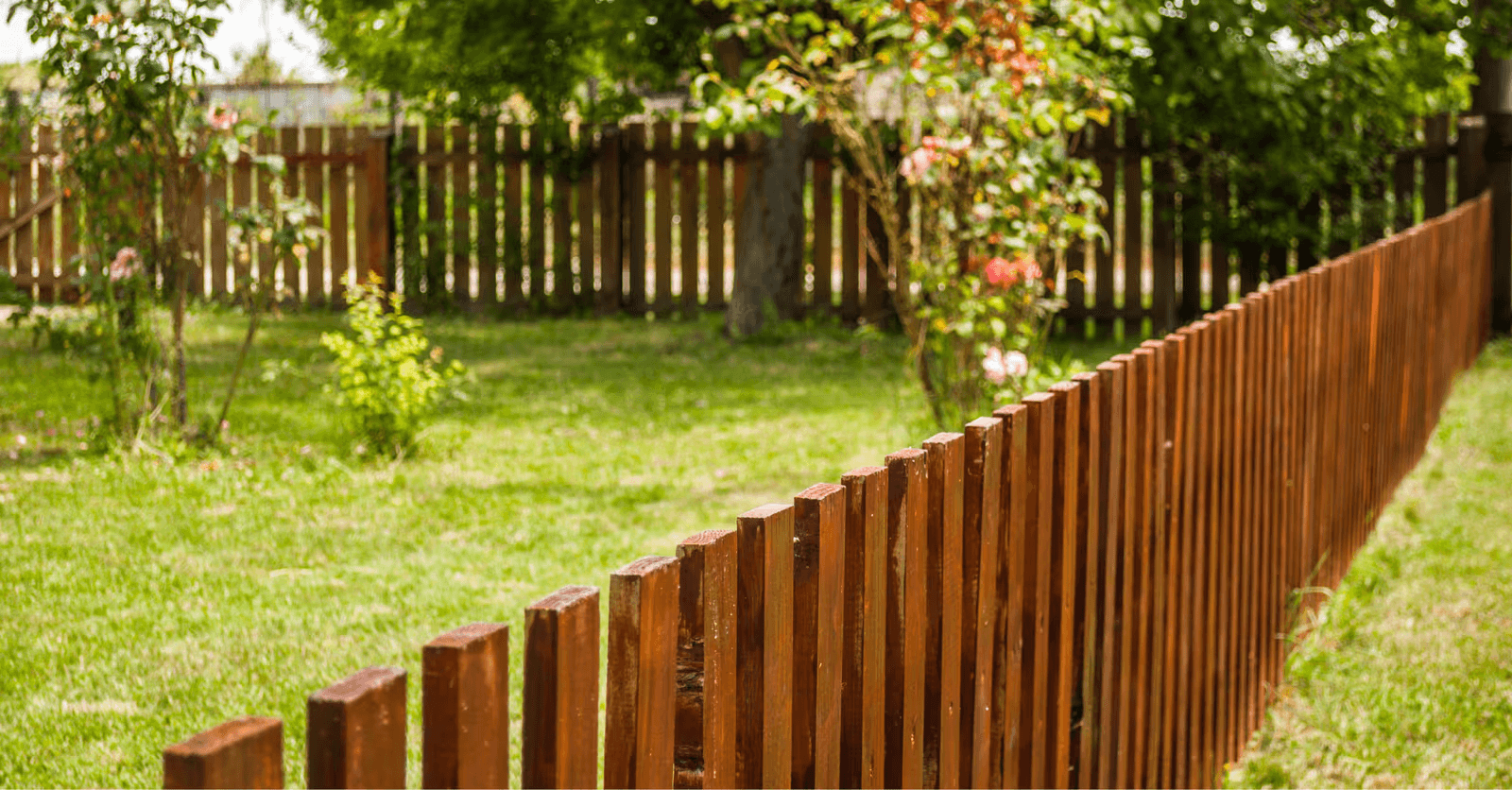 Wooden picket fence with vertical brown slats enclosing a lush green residential garden with grass, trees, and flowering shrubs.