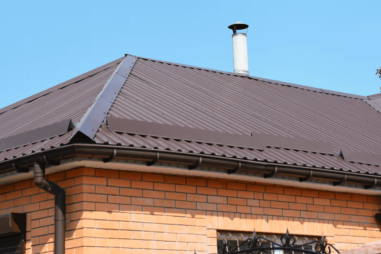 Residential brown corrugated metal roof with ribbed steel panels, ridge and hip flashing, metal chimney, gutters and downspouts on an orange brick house.