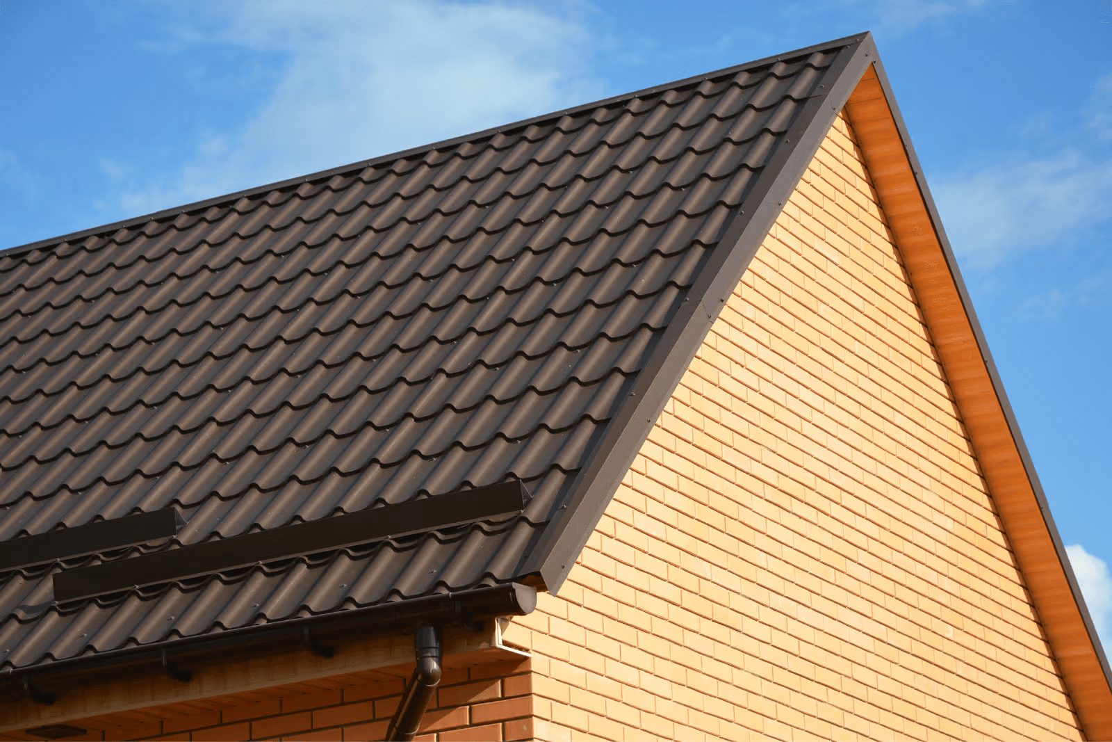 Residential roof with dark charcoal gray metal roofing tiles, snow guards, dark gutters, fascia trim and orange brick gable on a house with a pitched roof.