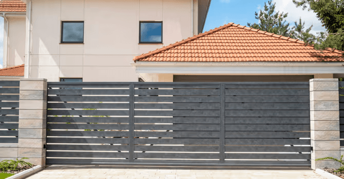 Modern gray composite driveway gate with horizontal slats and solid privacy panels, installed between stone pillars in front of a contemporary house with a terracotta tile roof.