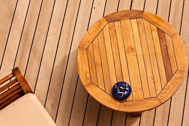 Overhead view of a light wood deck with a round natural wooden table, blue ashtray and outdoor chair with beige cushion.
